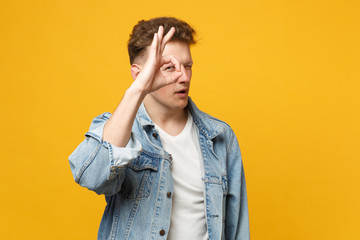 Suspicious young man in denim casual clothes holding hand near eyes imitating glasses or binoculars isolated on yellow orange background. People sincere emotions lifestyle concept. Mock up copy space.