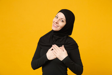 Smiling beautiful young arabian muslim woman in hijab black clothes put hands on heart isolated on yellow wall background studio portrait. People religious Islam lifestyle concept. Mock up copy space.