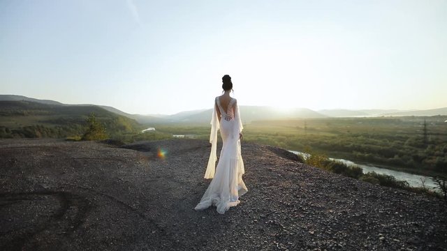 Young beautiful woman in wedding dress walks in mountains at sunset. Back view