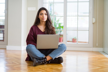 Young woman using computer laptop sitting on the floor with serious expression on face. Simple and...