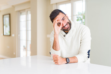 Handsome hispanic man wearing casual white sweater at home doing ok gesture with hand smiling, eye looking through fingers with happy face.