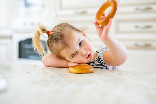 Little toddler kid having lunch in the warm sunny kitchen. Blonde girl with funny ponytail playing with two tasty bagels