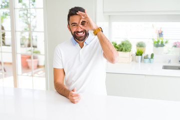 Handsome hispanic man casual white t-shirt at home doing ok gesture with hand smiling, eye looking through fingers with happy face.
