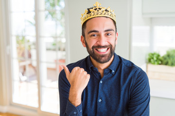 Handsome hispanic man wearing golden crown over head as the king smiling with happy face looking and pointing to the side with thumb up.