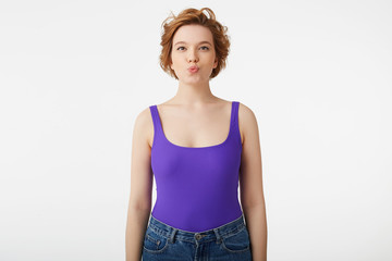 Portrait of young cute short-haired girl, wears in purple shirt and jeans, looks at the camera and keeps lips folded, stands over white background.