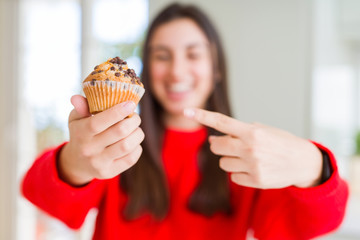 Beautiful young woman eating chocolate chips muffin very happy pointing with hand and finger