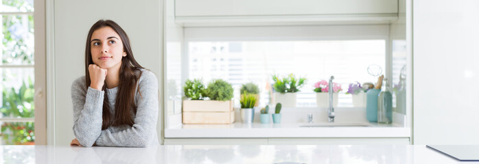 Wide angle picture of beautiful young woman sitting on white table at home with hand on chin thinking about question, pensive expression. Smiling with thoughtful face. Doubt concept.