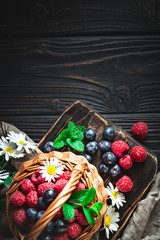 Raspberries and blueberries in a basket with chamomile and leaves on a dark background. Summer and healthy food concept. Selective focus. Background with copy space. Flat lay.