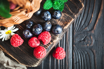 Raspberries and blueberries in a basket with chamomile and leaves on a dark background. Summer and healthy food concept. Selective focus. Flat lay.