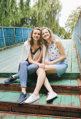 Two hipster girls in cool glasses are sitting on the old wooden bridge. Young fashionable sisters are having fun outdoors. Summer photoshoot for friends in the nature outdoors. Lifestyle concept.