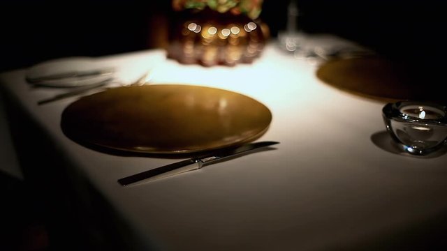 A butler wearing gloves, preparing the table for dinner by carefully adjusting (perfectly aligning) the cutlery and the glass. Dim lights.