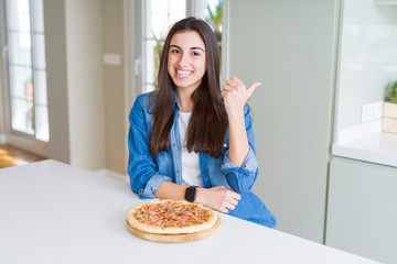 Beautiful young woman eating homemade tasty pizza at the kitchen smiling with happy face looking and pointing to the side with thumb up.