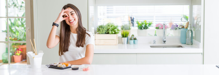 Wide angle picture of beautiful young woman eating asian sushi from delivery doing ok gesture with hand smiling, eye looking through fingers with happy face.