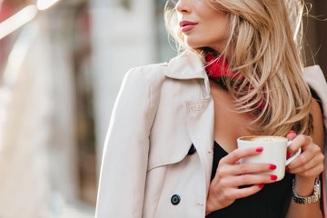 Close-up portrait of refined blonde woman in coat holding white cup with beverage on blur...
