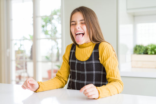 Young beautiful blonde kid girl wearing casual yellow sweater at home sticking tongue out happy with funny expression. Emotion concept.