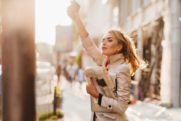 Outdoor portrait of active business-woman in trendy attire waiting for taxi in morning. Adorable fair-haired girl holding phone and documents looking away with inspired face expression. © Look!