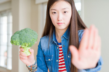 Beautiful Asian woman eating green fresh broccoli with open hand doing stop sign with serious and...