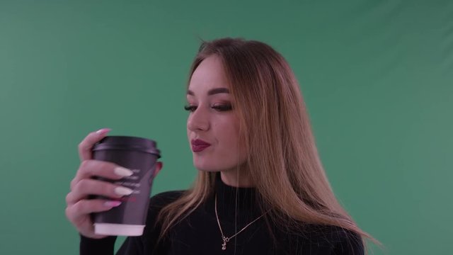 Young beautiful woman in black casual clothes is holdind a cup of coffee or tea in her hands and drinking standing in front of on green screen