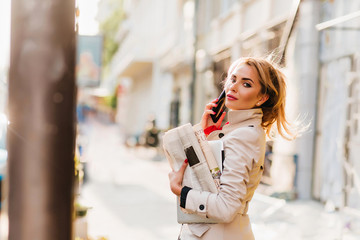 Adorable blonde business-woman enjoying city views while going to office with newspapers. Refined pretty girl in stylish light-brown coat looking back, calling someone.