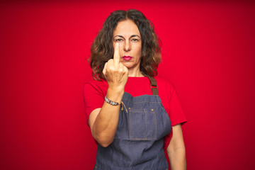 Middle age senior woman wearing apron uniform over red isolated background Showing middle finger, impolite and rude fuck off expression