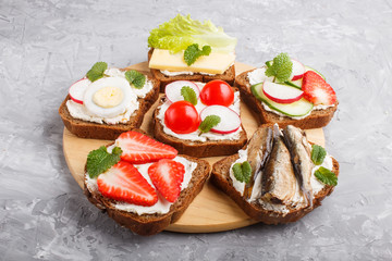 Set of different sandwiches with cheese, radish, lettuce, strawberry, sprats, tomatoes and cucumber on wooden board on a gray concrete background. side view.
