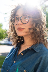 Outdoor close up portrait of young beautiful stylish happy smiling curly girl wearing sunglasses, posing in street. Sunny day light. Summer fashion concept. Copy, empty space for text