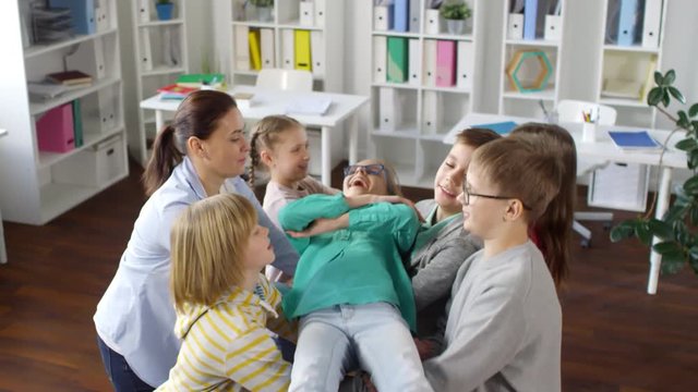 Waist-up shot of smiling teenage girl wearing turquoise top and glasses standing with closed eyes and falling backwards into other kids&rsquo; arms, during group training at female psychologist&rsquo;s office