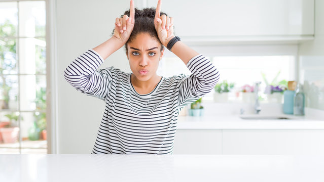 Beautiful african american woman with afro hair wearing casual striped sweater doing funny gesture with finger over head as bull horns
