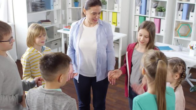 High-angle Medium Shot Of Casually Dressed Young Kids Standing In Circle With School Counselor In Her Sunny Office And Playing Brain Teaser Game Together, Watching Each Other Attentively And Laughing