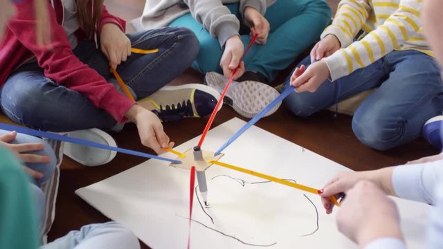 Tilting shot of group of kids sitting on floor together in circle and each holding colorful ribbon tied to marker in middle, trying to draw something with collective effort on white sheet of paper