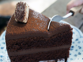 Cutting piece of chocolate fudge cake; decorated with cocoa powder, wafer and chocolate sauce, with fork served on white plate in wooden board. Selective focus.