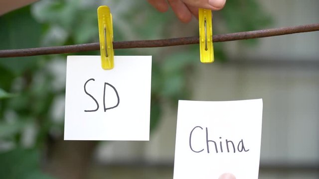 Inscriptions on white stickers on yellow clothespins on blurred background. Papers with the words SD on left and China on right hanging on metal rod with wind. Man hand hang word on right.