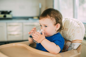 A little girl in the kitchen in the afternoon in a high chair drinking water from a glass glass