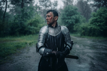 A young man stands in the image of a medieval knight in armor and with a sword.