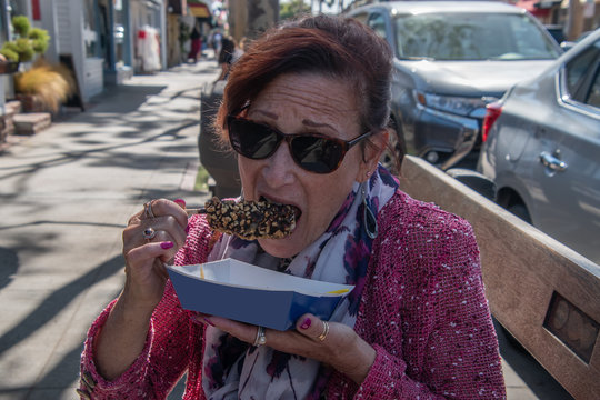 Middle Aged Pretty Baby Boomer Woman With Red Hair Red Sweater Colorful Scarf Sitting On A Bench Eating A Chocolate Covered Banana With Caramel And Crushed Nuts.