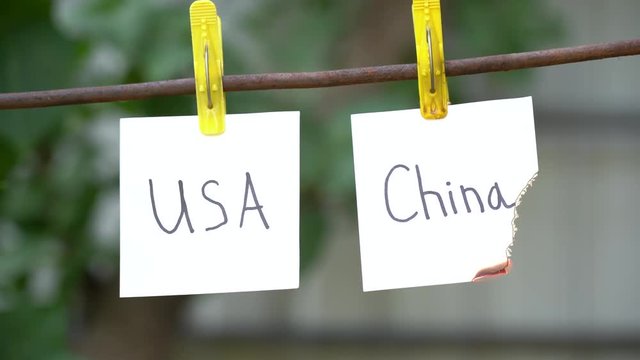 Inscriptions on white stickers on yellow clothespins on blurred background. Papers with words USA on left and China on right hanging on metal rod. Man&rsquo;s hand use red lighter ignites the word on right
