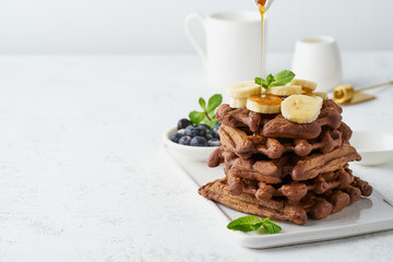 Chocolate banana waffles on white table, copy space, side view. Sweet brunch, maple syrup flow.