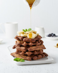 Chocolate banana waffles with maple syrup flow in milk jug, creamer on white table, side view,...