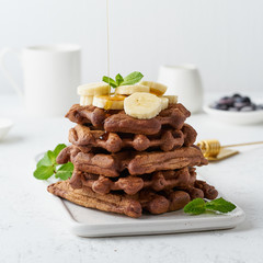 Chocolate banana waffles with maple syrup on white table, side view. Sweet brunch, maple syrup flow.