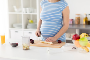 Obraz na płótnie Canvas cooking, pregnancy and healthy eating concept - pregnant woman with kitchen knife chopping fruits on wooden cutting board at home