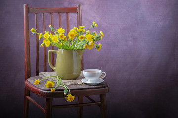A bouquet of dandelions in a vase and a cup of tea on a chair.
