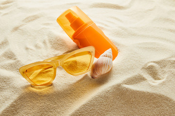 Fototapeta na wymiar yellow stylish sunglasses and sunscreen in orange bottle on sand with seashell