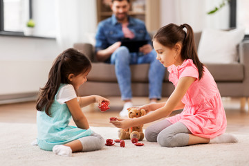 childhood, leisure and family concept - little sisters playing tea party game with toy crockery and teddy bear at home
