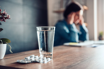Woman with headache and capsules with glass of water on a office desk - obrazy, fototapety, plakaty