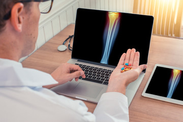 Doctor holding pills with x-ray of leg with pain on the knee on a laptop. Digital tablet on the wooden desk