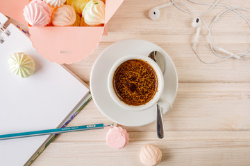Morning coffee with a notebook and a pencil, with a box filled with small meringues, headphones on a wooden light background.Топ view.
