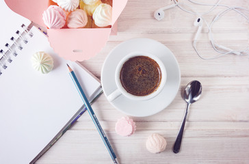 Morning coffee with a notebook and a pencil, with a box filled with small meringues, headphones on a wooden light background.Топ view.