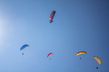 five paragliders on colored parachutes fly against the background of clean blue sky and the sun