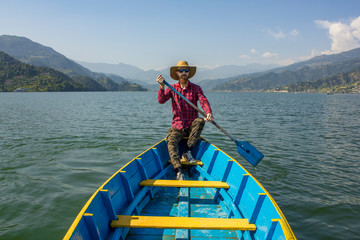 bearded man in a red shirt, hat and sunglasses in a blue wooden boat with a paddle in his hands, on the lake against the background of a green mountain valley