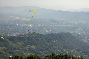 many paragliders on bright colored parachutes fly over the city in a foggy mountain valley aerial view
